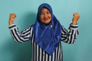 un emocionado de edad mediana asiático mujer en un azul hijab y a rayas camisa muestra un fuerte gesto por levantamiento su brazos y flexionando su músculos, sonriente con orgullo a el cámara, aislado en azul antecedentes. foto