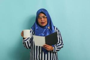 Calm Asian middle-aged woman, wearing a blue hijab, eyeglasses, and a striped shirt, holds an open book and a mug while standing against a blue background. photo