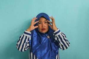 Middle-aged Asian woman, wearing a blue hijab and a striped shirt, tries to open her eyes with her fingers, feeling bored and tired from morning fatigue, while standing against a blue background photo