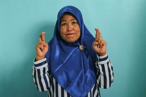 Portrait of a worried yet hopeful middle-aged Asian woman in hijab, shows her begging for something with fingers crossed, looking at camera and making a wish, while standing against a blue background photo