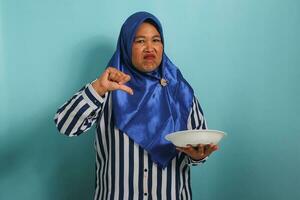 An angry middle-aged Asian woman, wearing a blue hijab and striped shirt, displays an unhappy expression, shows thumbs down, and holds a white plate, isolated against a blue background photo