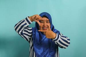un inspirado de edad mediana asiático mujer, en un azul hijab y a rayas camisa, hace un marco gesto, buscando el Perfecto ángulo o inspiración a capturar un momento en un foto. aislado en un azul antecedentes foto
