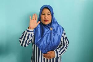un grave de edad mediana asiático mujer en un azul hijab y a rayas camisa es haciendo un jurar o promesa gesto mientras mirando a el cámara. ella es aislado terminado un azul antecedentes. foto