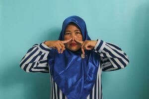 A middle-aged Asian woman, in a blue hijab, holds her breath and pinches her nose with her fingers, reacting to a stinky and disgusting, intolerable bad smell, while standing against a blue background photo