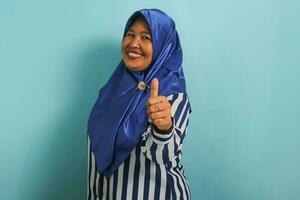 An excited middle-aged Asian woman in a blue hijab and striped shirt shows a thumbs-up and an okay sign, symbolizing success and expressing approval, while standing against a blue background. photo