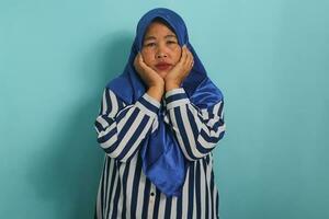 Bored middle-aged Asian woman, wearing a blue hijab and a striped shirt, appears unhappy or tired, dealing with problems and possibly experiencing depression, while standing against a blue background. photo