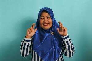 An excited middle-aged Asian woman, wearing a blue hijab, is hopeful and optimistic, crossing her fingers for good luck, making a wish, and smiling while standing against a blue background photo