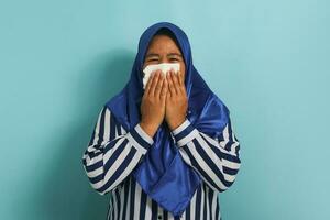 insalubre de edad mediana asiático mujer, vistiendo un azul hijab y un a rayas camisa, es visto soplo su líquido nariz con un pañuelo de papel mientras estornudos debido a el gripe. cuidado de la salud y médico concepto foto