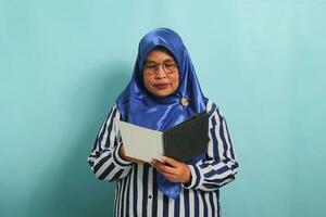 Asian middle-aged woman, wearing a blue hijab, eyeglasses, and a striped shirt, holds an open book while standing against a blue background. photo