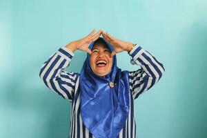 An excited middle-aged Asian woman in a blue hijab and striped shirt is making a home or roof gesture by holding her hands above her head while standing against a blue background photo