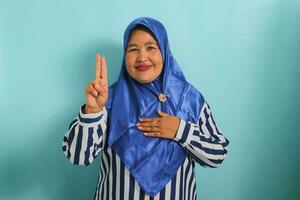 A Confident middle-aged Asian woman in a blue hijab and striped shirt is making a swearing or promise gesture while looking at the camera with hand on chest. She is isolated over a blue background. photo
