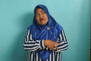 An upset middle-aged Asian woman in a blue hijab and striped shirt stands with her arms folded, feeling offended, unsatisfied, disappointed, displeased, moody, and jealous, isolated on blue background photo