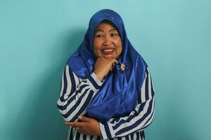 A displeased middle-aged Asian woman, wearing a blue hijab and a striped shirt, is displaying a disgusted expression while repulsed by something, while standing against a blue background photo
