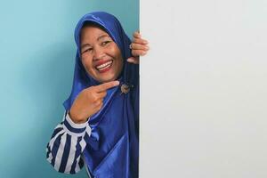 Excited middle-aged Asian woman in a blue hijab and striped shirt holds and pointing a blank white banner signboard with copyspace, offering ample space for your message, against a blue background. photo