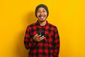 Asian man in a plaid shirt and beanie hat holding a mobile phone isolated on yellow background. photo