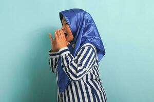 An excited middle-aged Asian woman in a blue hijab and striped shirt shouts and announce something while standing against a blue background. photo