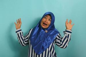 A scared middle-aged Asian woman in a blue hijab and striped shirt gasps and jumps in fear while standing against a blue background. photo