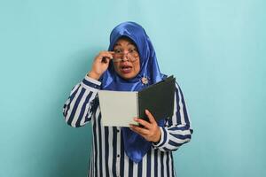Amazed Asian middle-aged woman, wearing a blue hijab, eyeglasses, and a striped shirt, holds an open book with a surprised expression while standing against a blue background. photo