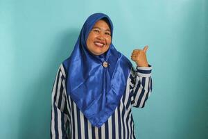 An excited middle-aged Asian woman in a blue hijab and striped shirt points to the left with her thumb, highlighting the copyspace or empty space, isolated over blue background. photo