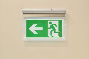 Close-up of green emergency exit sign showing the way to escape on wall. Fire exit in corridor of building. Fire prevention and house safety concept. Arrow pointing exit photo