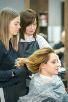 dos hembra peluqueros preparar largo pelo de un joven mujer haciendo chinos peinado en un belleza salón. foto