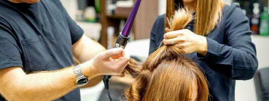 dos peluqueros utilizando curling hierro en clientes largo marrón pelo en un belleza salón. foto