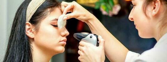 Side view of a make-up artist using aerograph making an airbrush makeup foundation on a female face in a beauty salon. photo