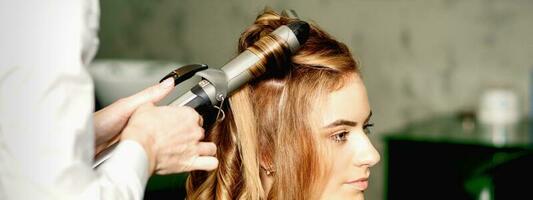 Hairdresser using curling tongs curls long brown hair on the young caucasian girl in a beauty salon. photo