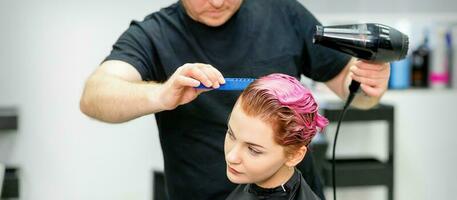 A hairdresser is drying the pink hair of the young woman in a beauty salon. photo