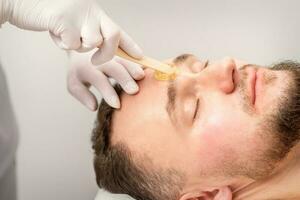 cosmetólogo aplicando cera pegar Entre Cejas durante el procedimiento de depilación en el belleza salón. foto