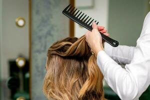 un hembra peluquero es peinada el largo marrón pelo de un joven mujer a un salón. foto