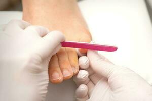 Pedicurist hands in protective rubber gloves filing toenails on feet with a nail file in a beauty salon. photo