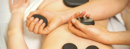 Hot stone massage on the female back with hands of masseur holding black massage stones in spa salon. photo