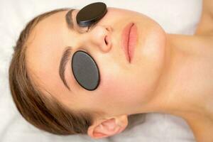 Young woman receiving facial massage with black mineral stones on her eyes in a spa. photo