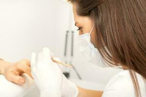 Manicure master in protective face mask and white gloves apply polish to female fingernails. photo