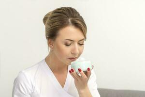 Portrait of young beautician sniffing moisturizing cream holding white jar on white background, copy space. photo