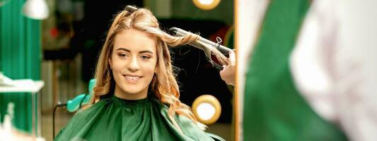 Hairdresser using curling tongs curls long brown hair on the young caucasian girl in a beauty salon. photo