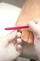 Pedicurist hands in protective rubber gloves filing toenails on feet with a nail file in a beauty salon. photo