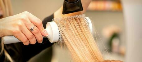 Professional hairdresser dry hair with a hairdryer and round hairbrush in a beauty salon. photo