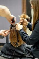 The rear view of two hairdressers are curling hair for a young woman with electric hair iron in a beauty salon. photo