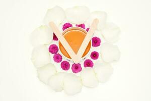 Jar of wax with wooden sticks, pink flowers, and white petals on white background. Concept of depilation. Flat lay top view. photo