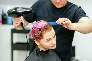 un peluquero es el secado el rosado pelo de el joven mujer en un belleza salón. foto