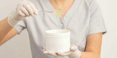 Liquid yellow sugar paste for depilation on a stick flows into the jar in hands of a beautician. photo