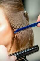 Hairdresser straightens female brown hair of medium length with a hair with an iron hair straightener and comb in a beauty salon. photo