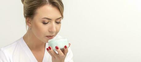 Portrait of young beautician sniffing moisturizing cream holding white jar on white background, copy space. photo