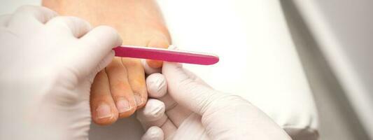 Pedicurist hands in protective rubber gloves filing toenails on feet with a nail file in a beauty salon. photo