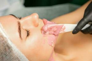 Beautician applying alginate peel-off powder facial mask with the spatula in a spa. photo