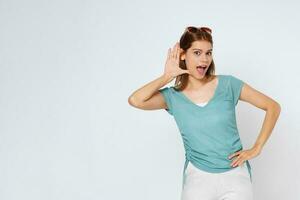 Portrait of young woman with opened mouth trying to hear something with raised hand near her ear and looking at camera, while standing over white isolated background. photo