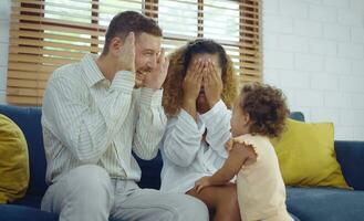 Happy cheerful parents and cute daughter are playing peekaboo on sofa together. photo