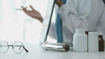 Medical worker in lab coat and sterile mask, doing a microscope analysis while her colleague are working behind video